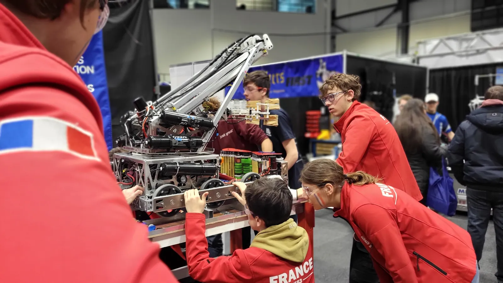 Des élèves en veste rouge France penchés sur leur robot pour l'ajuster, dans les stands d'une compétition