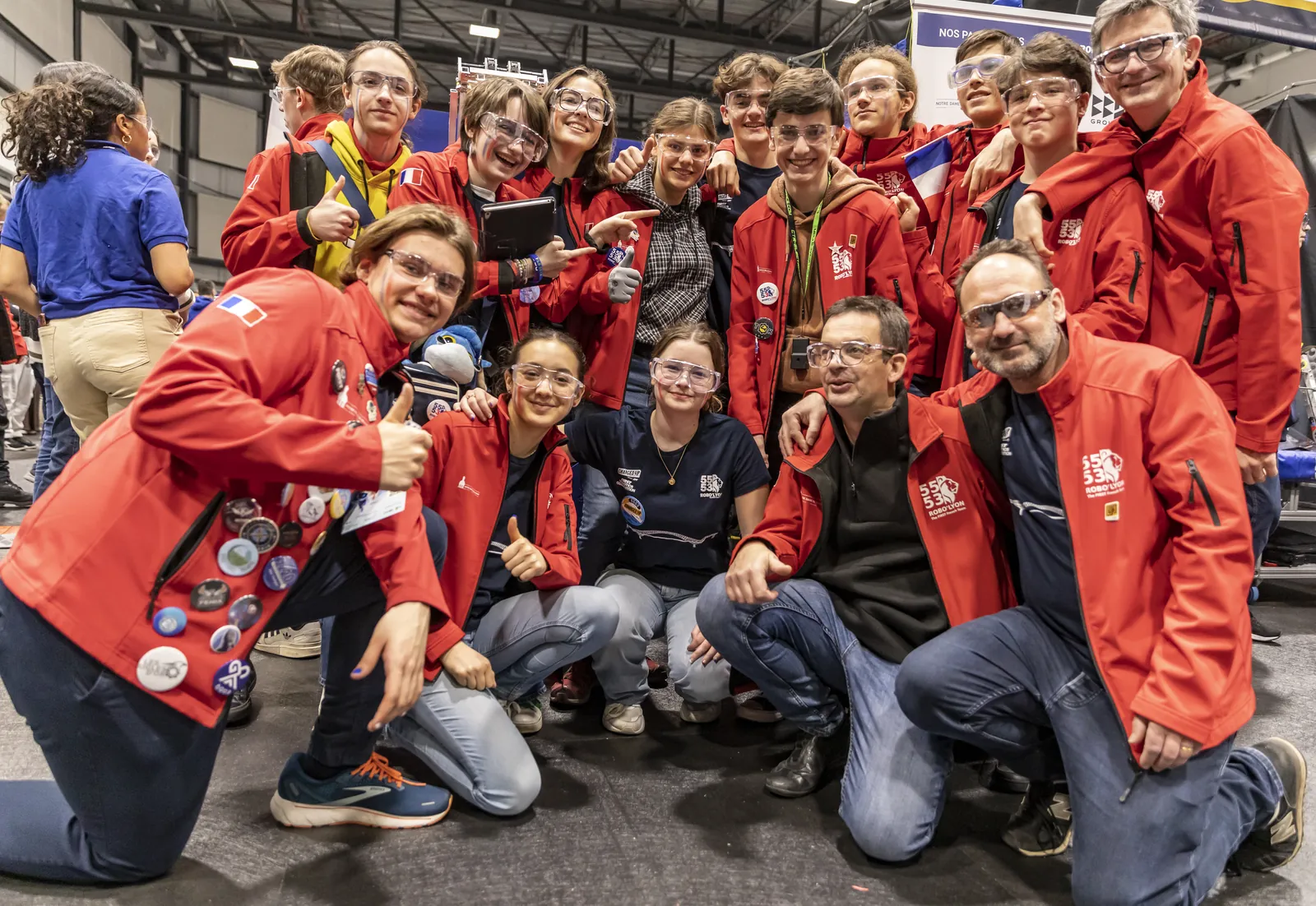 L'équipe 5553 au complet, élèves et mentors en veste rouge et lunettes de sécurité, pose en souriant dans les stands d'une compétition
