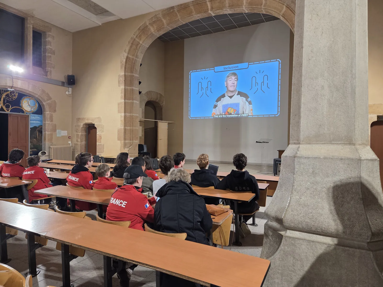Élèves en veste rouge France assis dans un amphithéâtre, face à la vidéo de lancement de saison FIRST projetée au mur