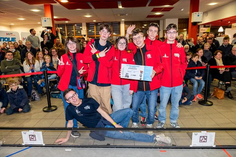 Une équipe Robo'Lyon en veste rouge pose avec son ticket pour la finale, devant le public de la régionale du 18 janvier